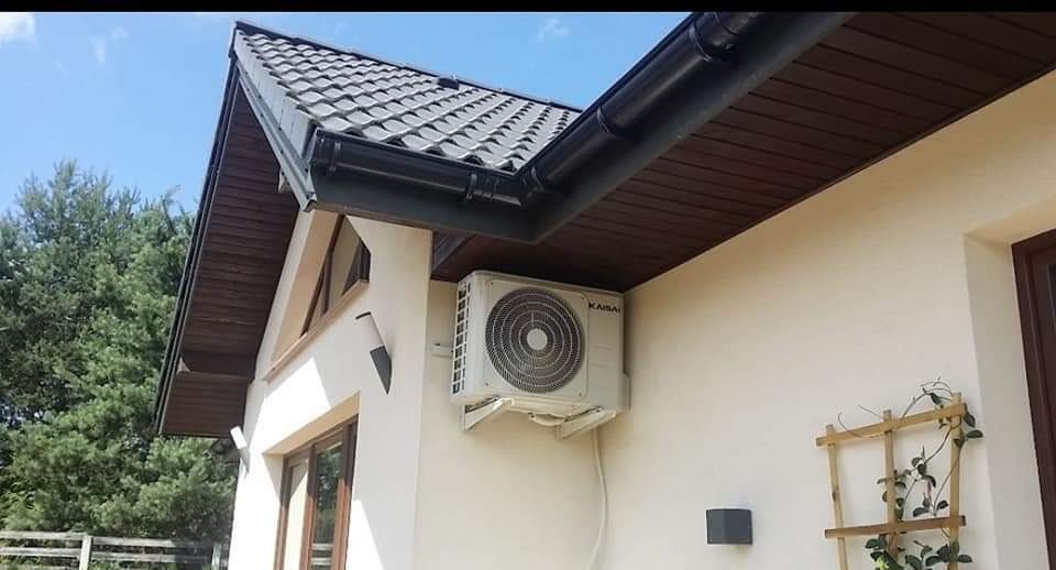 Zewnętrzna jednostka klimatyzacji marki Kaisai zamontowana na kremowej ścianie domu z ciemnobrązowym dachem i rynnami.