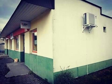 Zewnętrzna jednostka klimatyzacyjna zamontowana na ścianie budynku z zielonym cokołem i oknami z czerwonymi ramami, widok z boku, pochmurne niebo.