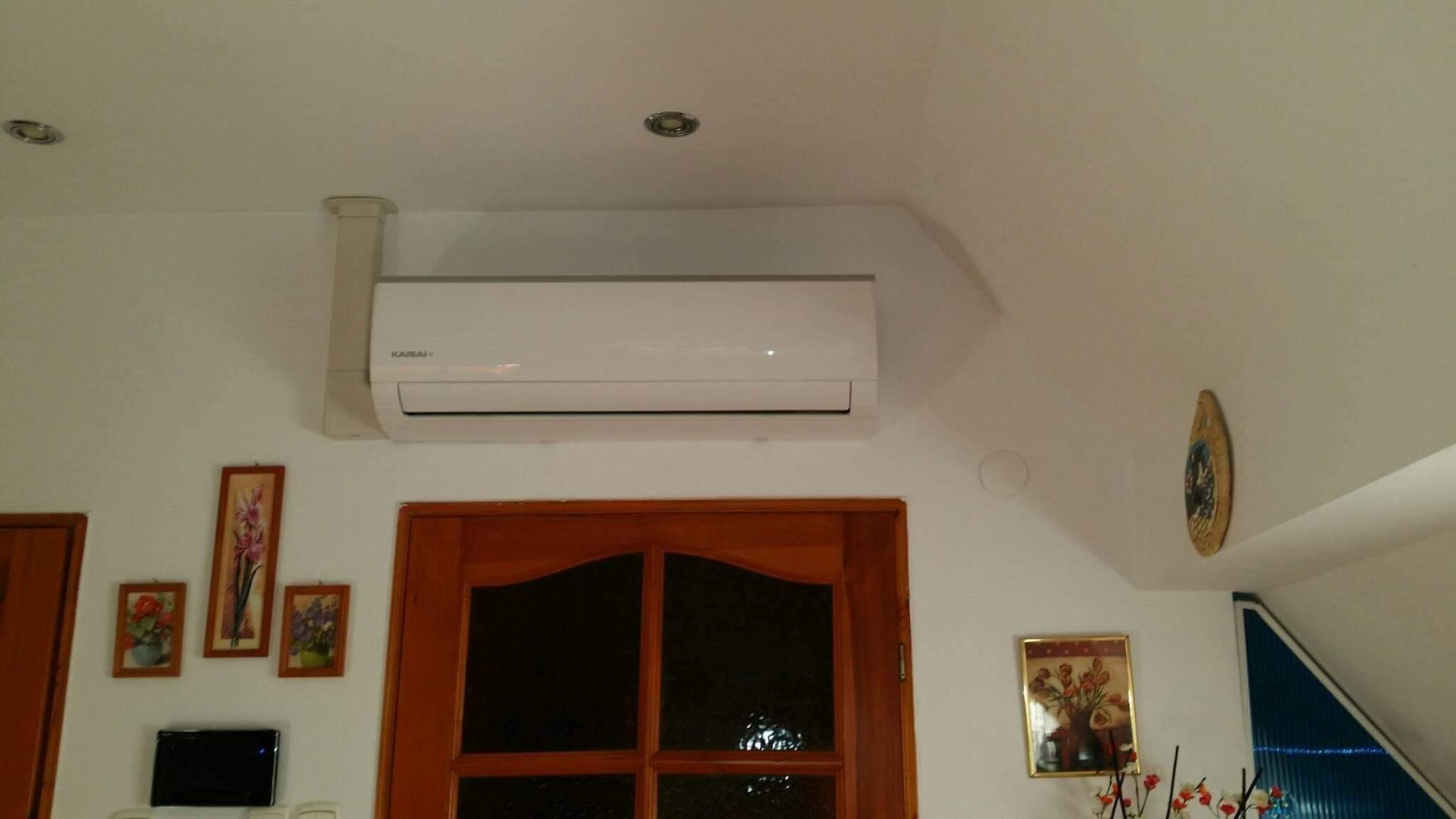 Biała jednostka wewnętrzna klimatyzatora marki Kaisai zamontowana na białej ścianie w pomieszczeniu z drewnianymi drzwiami i obrazami.