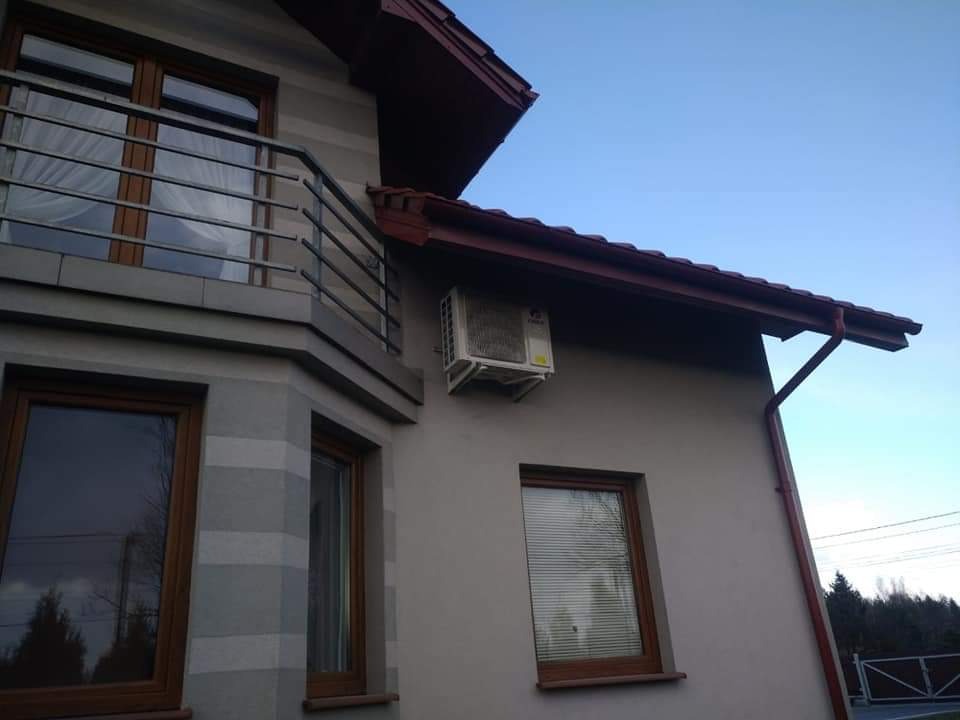 Biała jednostka zewnętrzna klimatyzatora zamontowana na szarej ścianie domu z brązowymi oknami i dachem, widok z dołu na błękitne niebo.