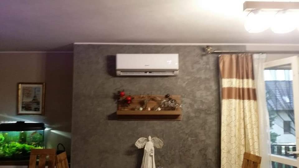 Wnętrze pokoju z zamontowanym, białym klimatyzatorem ściennym nad drewnianą półką z dekoracjami, na tle szarej ściany z fakturą betonu. Akwarium z rybkami widoczne po lewej stronie.