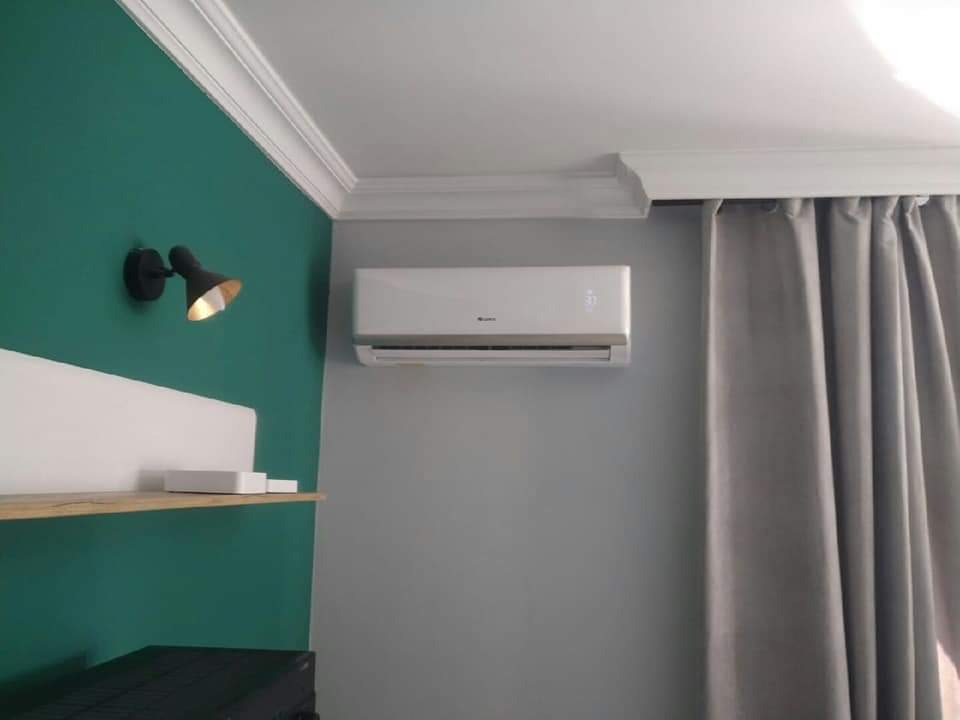 Nowoczesny biały klimatyzator ścienny zamontowany w szarym pokoju z zasłonami, widoczny fragment zielonej ściany z czarną lampą i półką.