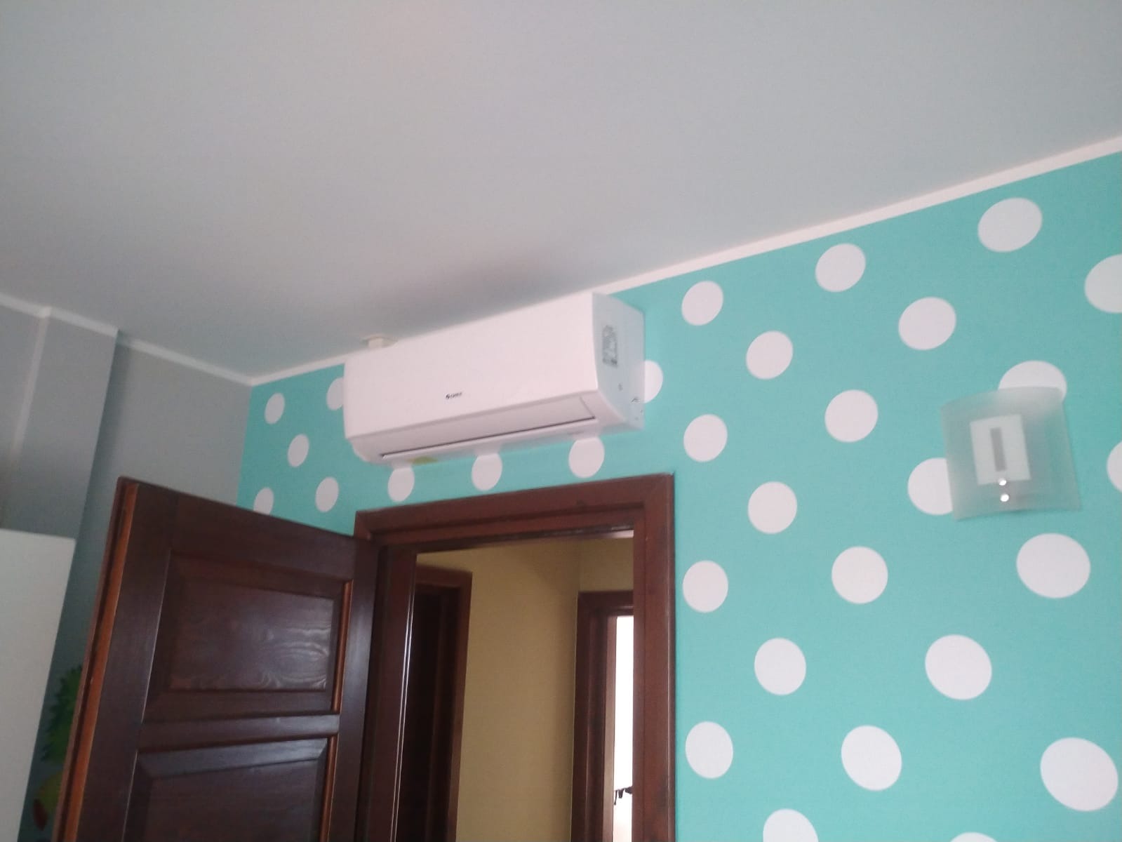 Biała jednostka wewnętrzna klimatyzatora marki Gree zamontowana na turkusowej ścianie w pokoju z białymi kropkami, widoczna nad brązową futryną drzwi.
