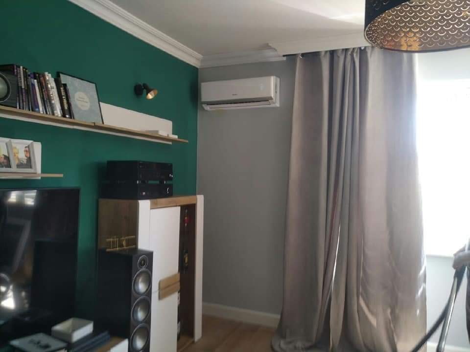 Biała klimatyzacja ścienna zamontowana w nowoczesnym salonie z zieloną ścianą, szarą ścianą, zasłonami i drewnianą podłogą.