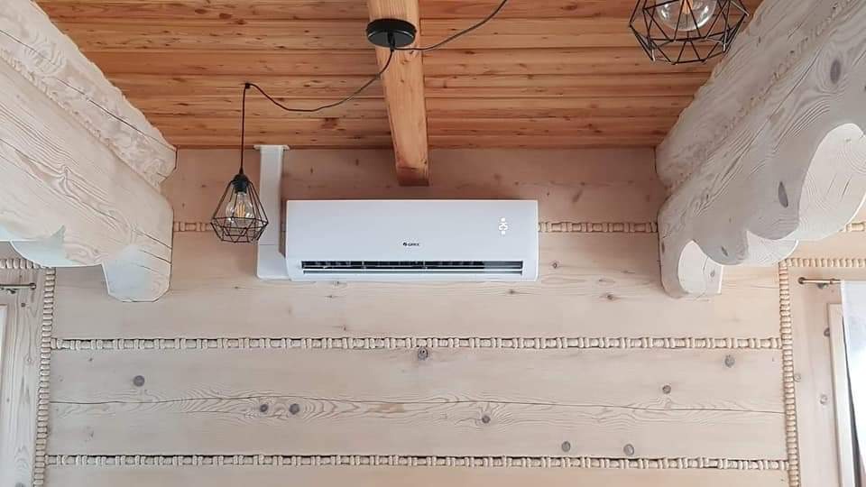 Biały klimatyzator marki Gree zamontowany na ścianie z jasnego drewna w stylu góralskim, widoczne drewniane belki stropowe i ozdobne lampy wiszące.