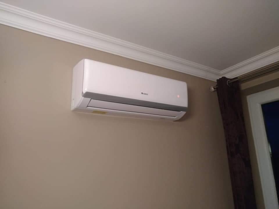 Biały klimatyzator marki Gree zamontowany na beżowej ścianie w pomieszczeniu z listwami dekoracyjnymi pod sufitem i zasłoniętym oknem.