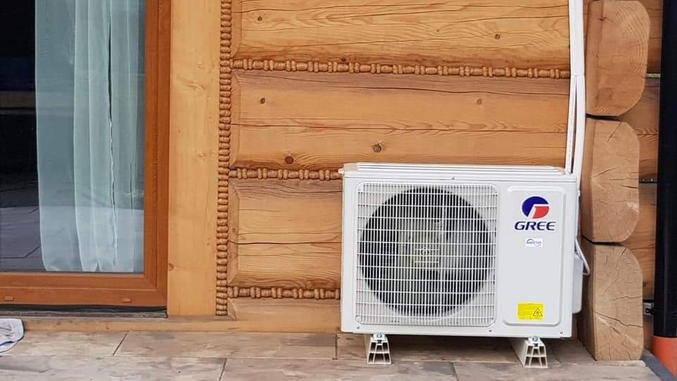 Biała jednostka zewnętrzna klimatyzatora marki Gree zamontowana na betonowej podstawie przy drewnianej ścianie domu z bali, widoczny fragment okna po lewej stronie.