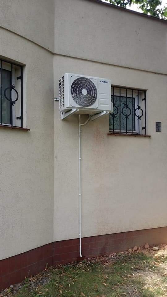 Zewnętrzna jednostka klimatyzatora Kaisai zamontowana na ścianie budynku, widoczne rury odprowadzające skropliny, okna z ozdobnymi kratami w tle.