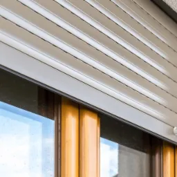 Zasłonięte okno z beżową roletą zewnętrzną, widoczny fragment elewacji i ramy okiennej.