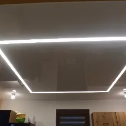 Błyszczący, szary sufit napinany z wbudowanym kwadratowym oświetleniem LED, widoczne dodatkowe punktowe lampy i odbicie wyposażenia kuchni.