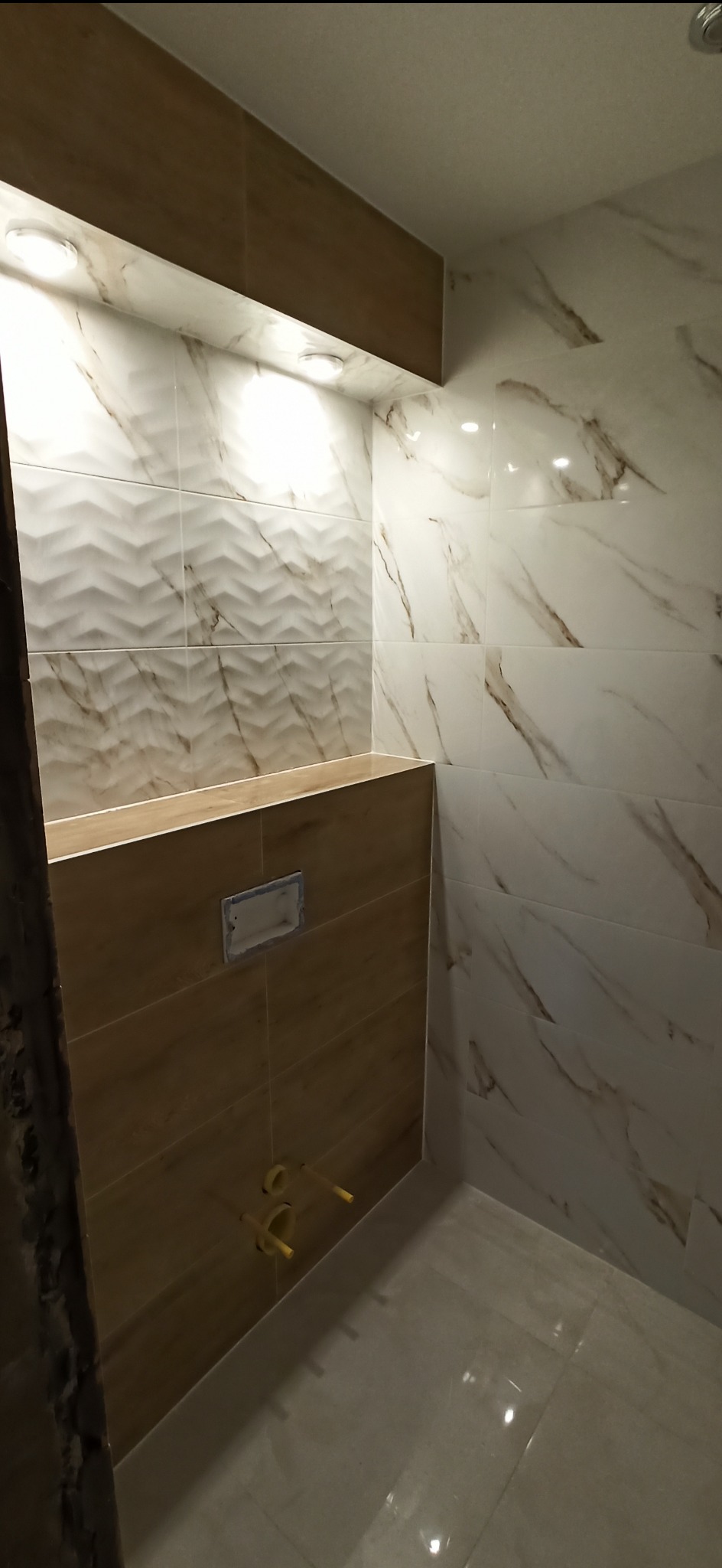 Wykończona łazienka z płytkami imitującymi marmur na ścianach i podłodze, oraz zabudową stelaża WC wykończoną płytkami drewnopodobnymi, widoczne instalacje wodne.