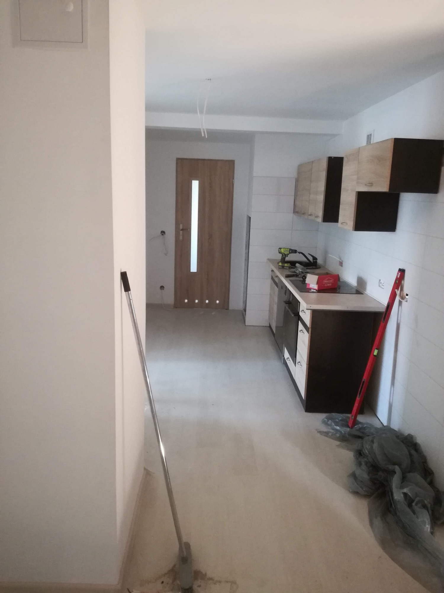 Wnętrze w trakcie prac wykończeniowych: widok na nową zabudowę kuchenną z czarno-drewnianymi szafkami, białe płytki na ścianie, drzwi wewnętrzne z przeszkleniem i narzędzia budowlane w tle.