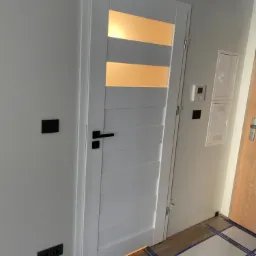 Białe drzwi wewnętrzne z poziomymi przeszkleniami, czarna klamka, szare ściany z czarnymi gniazdkami i włącznikami, zabezpieczona folią podłoga z paneli.