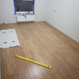 Fragment pokoju z ułożonymi panelami podłogowymi w odcieniach brązu, poziomica leży na podłodze, okno zabezpieczone folią malarską, białe ściany i grzejnik.