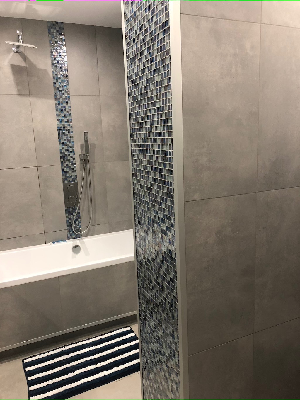 Nowoczesna łazienka wykończona szarymi płytkami wielkoformatowymi i mozaiką w odcieniach niebieskiego, z białą wanną i paskowanym dywanikiem podłogowym.