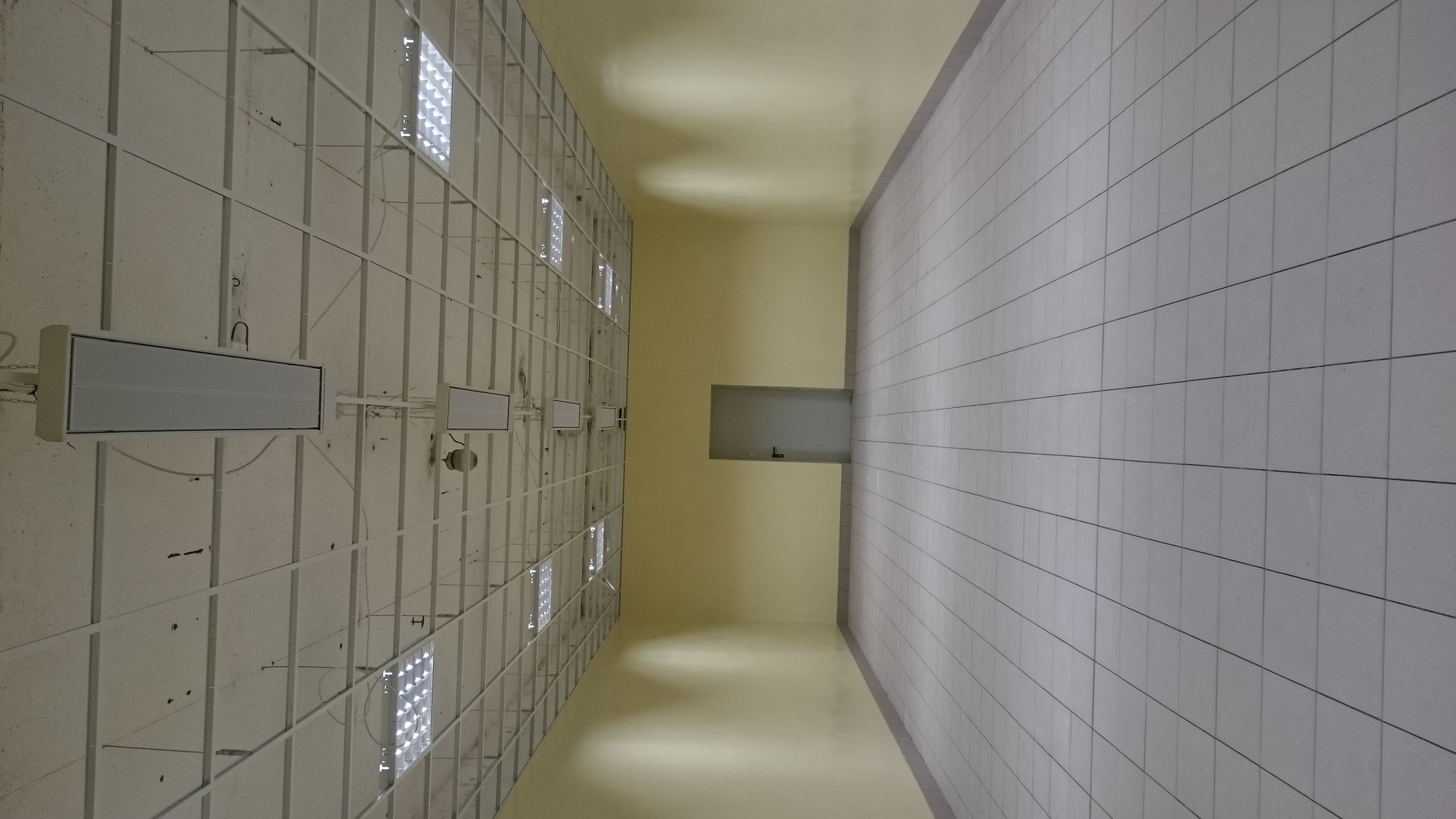 Wnętrze pomieszczenia z widoczną instalacją oświetleniową rastrową na suficie i fragmentem ściany wyłożonej jasnymi, kwadratowymi płytkami ceramicznymi.