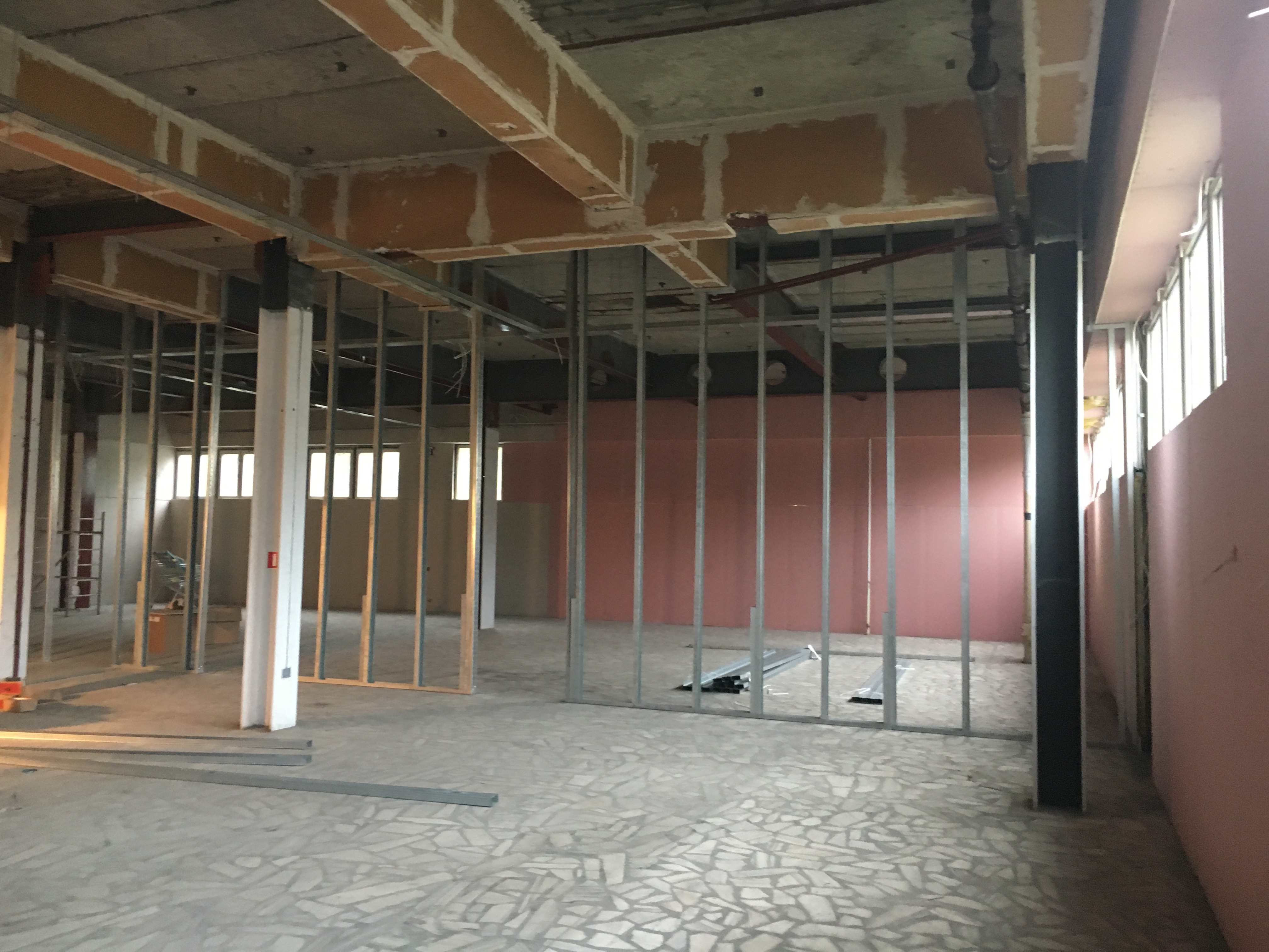 Wnętrze w trakcie prac: widoczne metalowe profile ścian działowych, fragmenty instalacji pod sufitem, surowy betonowy strop, mozaika na podłodze, ściany w odcieniach beżu i różu, okna w tle.