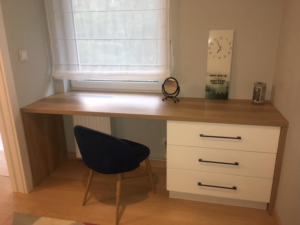 Drewniane biurko z białymi szufladami i granatowym krzesłem na drewnianej podłodze, z dekoracjami na blacie: małe lusterko, zegar z napisem 'never give up', świeca.