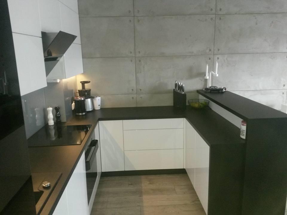 Nowoczesna kuchnia z białymi szafkami i czarnym blatem, betonowa ściana w tle, widoczne akcesoria kuchenne i dekoracje.