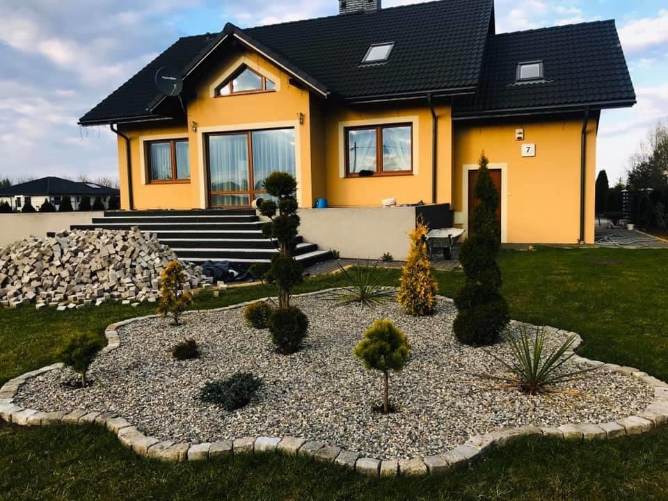 Ogród przed domem z nasadzeniami drzewek iglastych i ozdobnych na żwirowym podłożu, obrzeże z kostki brukowej, na trawniku stos kamieni, dom w tle.