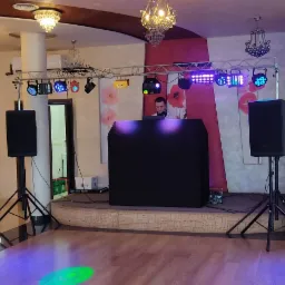 Sala bankietowa z przygotowanym stanowiskiem DJ-a: czarny podświetlany panel, głośniki na statywach, oświetlenie sceniczne na metalowej konstrukcji. W tle nakryte stoły.