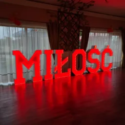 Duże, podświetlane na czerwono litery układające się w słowo 'MIŁOŚĆ' na drewnianej podłodze w sali z oknami zasłoniętymi białymi firankami.