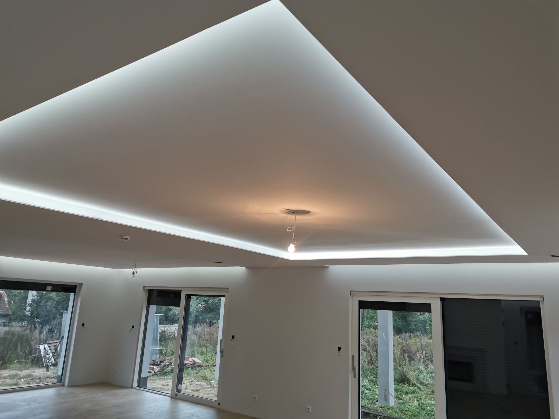 Nowoczesne wnętrze z podwieszanym sufitem oświetlonym ledami, widoczne duże okna przesuwne i surowa podłoga.