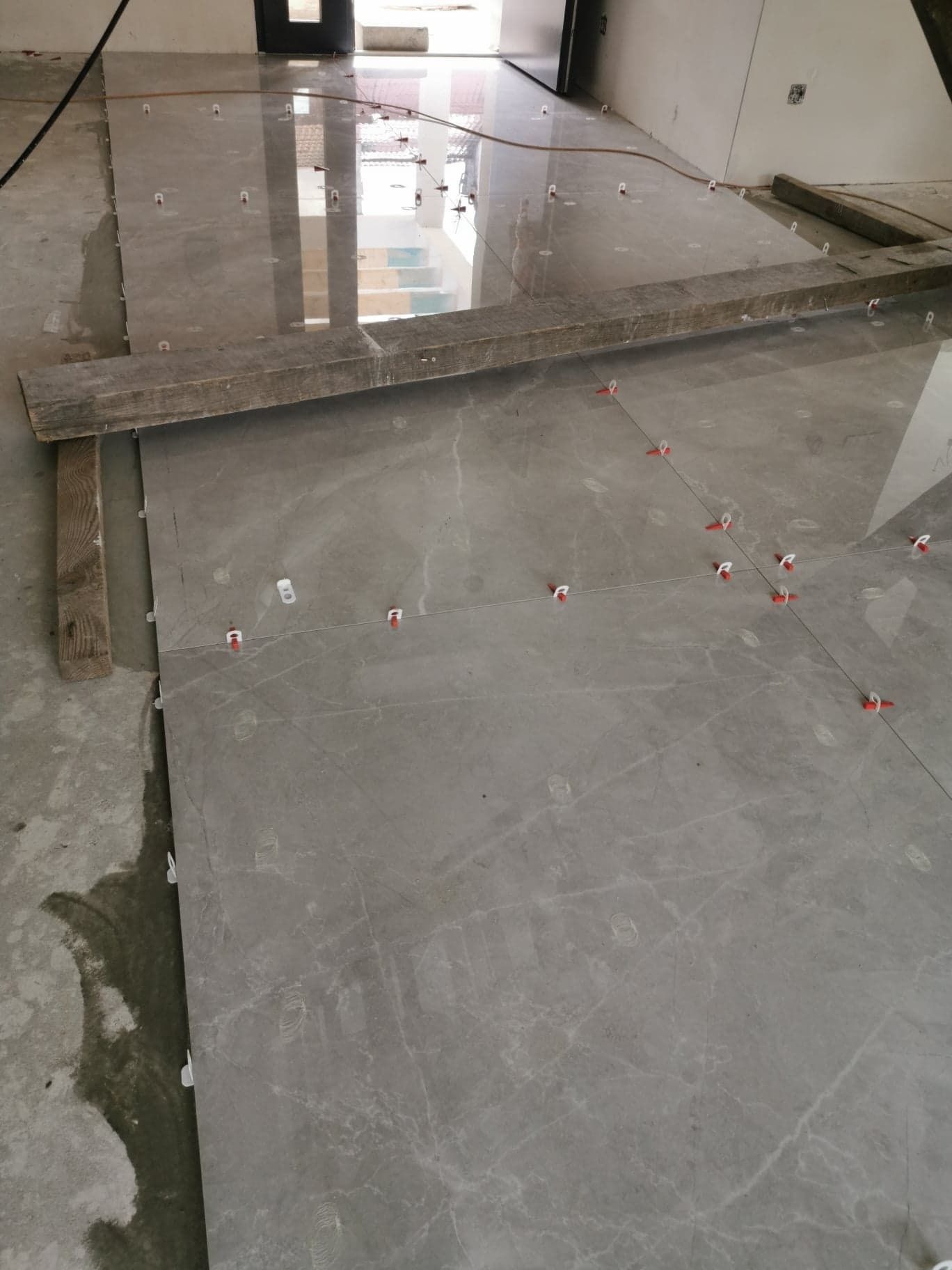 Układanie szarych płytek podłogowych z użyciem systemu poziomowania, widoczne deski pomocnicze i ślady wilgoci na surowym podłożu.