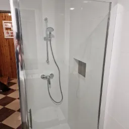 Szklana kabina prysznicowa z chromowaną armaturą, wnęką wykończoną drewnopodobną płytką oraz widoczną podłogą w szachownicę poza kabiną.