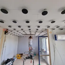 Montaż stelaża sufitu podwieszanego: widok na pracownika na drabinie mocującego elementy konstrukcji, z widocznym laserowym wyznacznikiem poziomu na ścianie i przygotowanym klimatyzatorem...