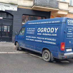 Dobema Niepowtarzalne Ogrody inż. Mariusz Wanzke - Niebieski van Volkswagen Transporter firmy DOBEMA OGRODY, reklamujący usługi ogrodnicze, w tym pielęgnację zieleni i systemy nawadniania, zaparkowany przy kamienicy.