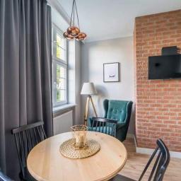 Art-Remo - usługi remontowo-budowlane - Jasne wnętrze pokoju z okrągłym drewnianym stołem, czarnymi krzesłami, szarą zasłoną, lampą na trójnogu, turkusowym fotelem i ścianą z czerwonej cegły z telewizorem.