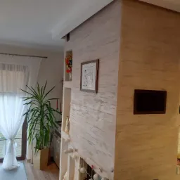 Ściana wewnętrzna z dekoracyjnym tynkiem w odcieniach beżu i szarości, z wnękami zawierającymi dekoracje, obrazek w ramce i roślinę doniczkową przy oknie z białymi zasłonami.