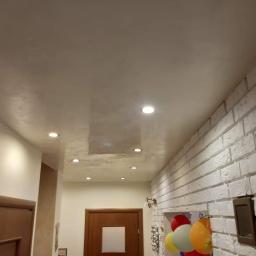 Art-Remo - usługi remontowo-budowlane - Wykończone wnętrze z dekoracyjnym tynkiem na suficie, oświetleniem punktowym i białą ścianą z cegły, widoczne drzwi i dekoracje wiszące na ścianie.