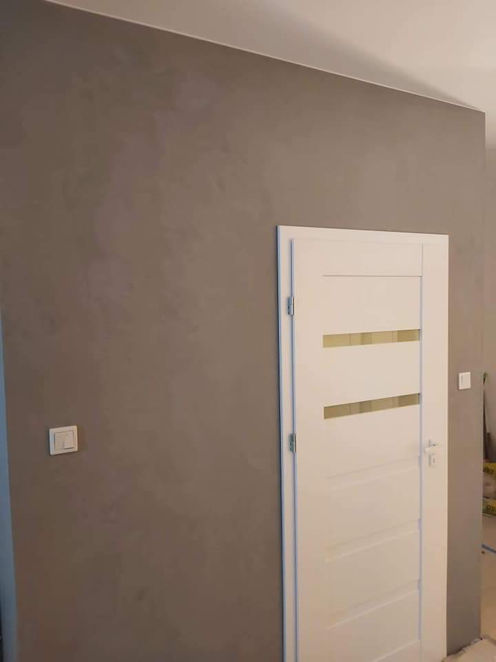 Ściana pomalowana farbą dekoracyjną w odcieniach szarości z efektem subtelnych przetarć, obok białe drzwi wewnętrzne z poziomymi przeszkleniami i chromowanymi okuciami, widoczny włącznik światła.