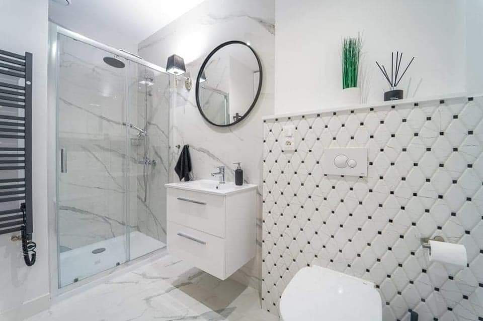 Nowoczesna łazienka z kabiną prysznicową, umywalką z szafką, okrągłym lustrem i biało-czarną mozaiką na ścianie za toaletą.
