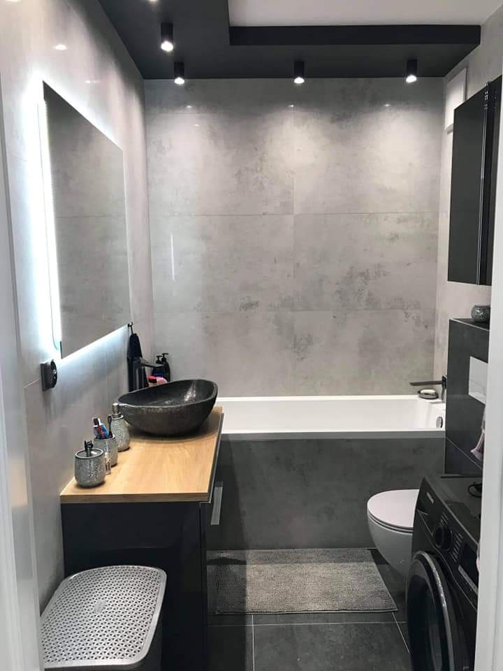 Nowoczesna łazienka z szarą, teksturowaną płytką, podświetlanym lustrem, umywalką w kształcie misy na drewnianym blacie, czarnym sufitem podwieszanym z punktowymi lampami i pralką w rogu.