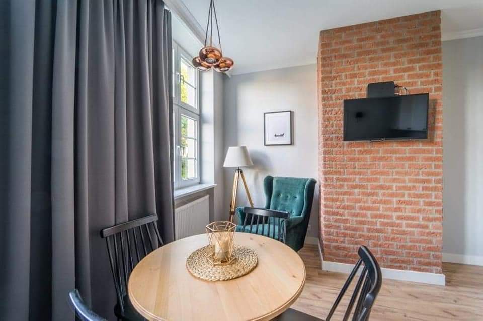 Jasne wnętrze pokoju z okrągłym drewnianym stołem, czarnymi krzesłami, szarą zasłoną, lampą na trójnogu, turkusowym fotelem i ścianą z czerwonej cegły z telewizorem.