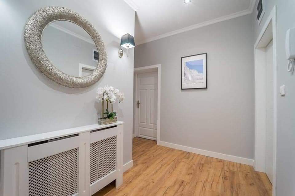 Jasny przedpokój z panelami podłogowymi imitującymi drewno, srebrnym lustrem w ozdobnej ramie, białym grzejnikiem z ażurową obudową i dekoracyjną białą orchideą w doniczce.