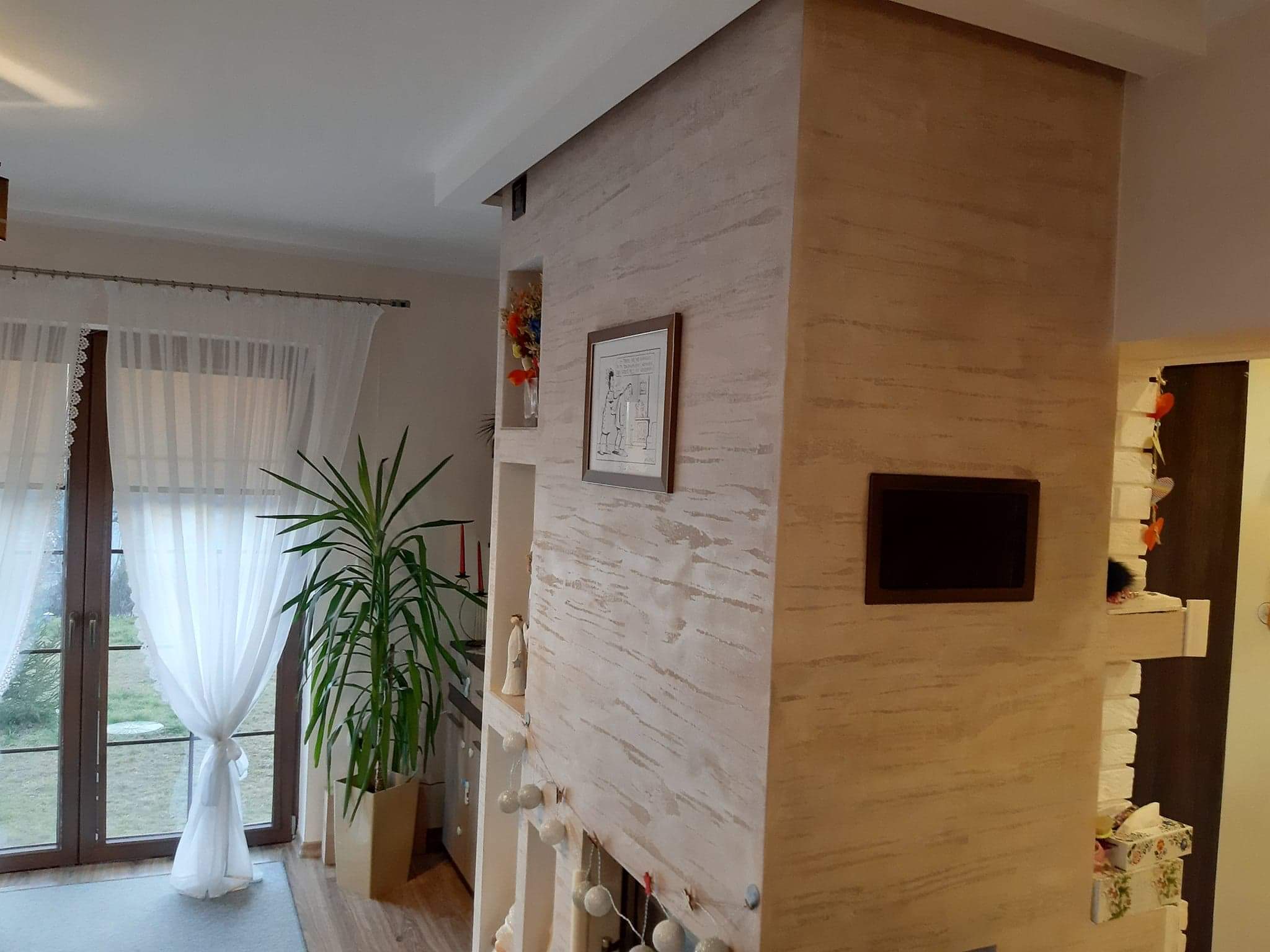 Ściana wewnętrzna z dekoracyjnym tynkiem w odcieniach beżu i szarości, z wnękami zawierającymi dekoracje, obrazek w ramce i roślinę doniczkową przy oknie z białymi zasłonami.