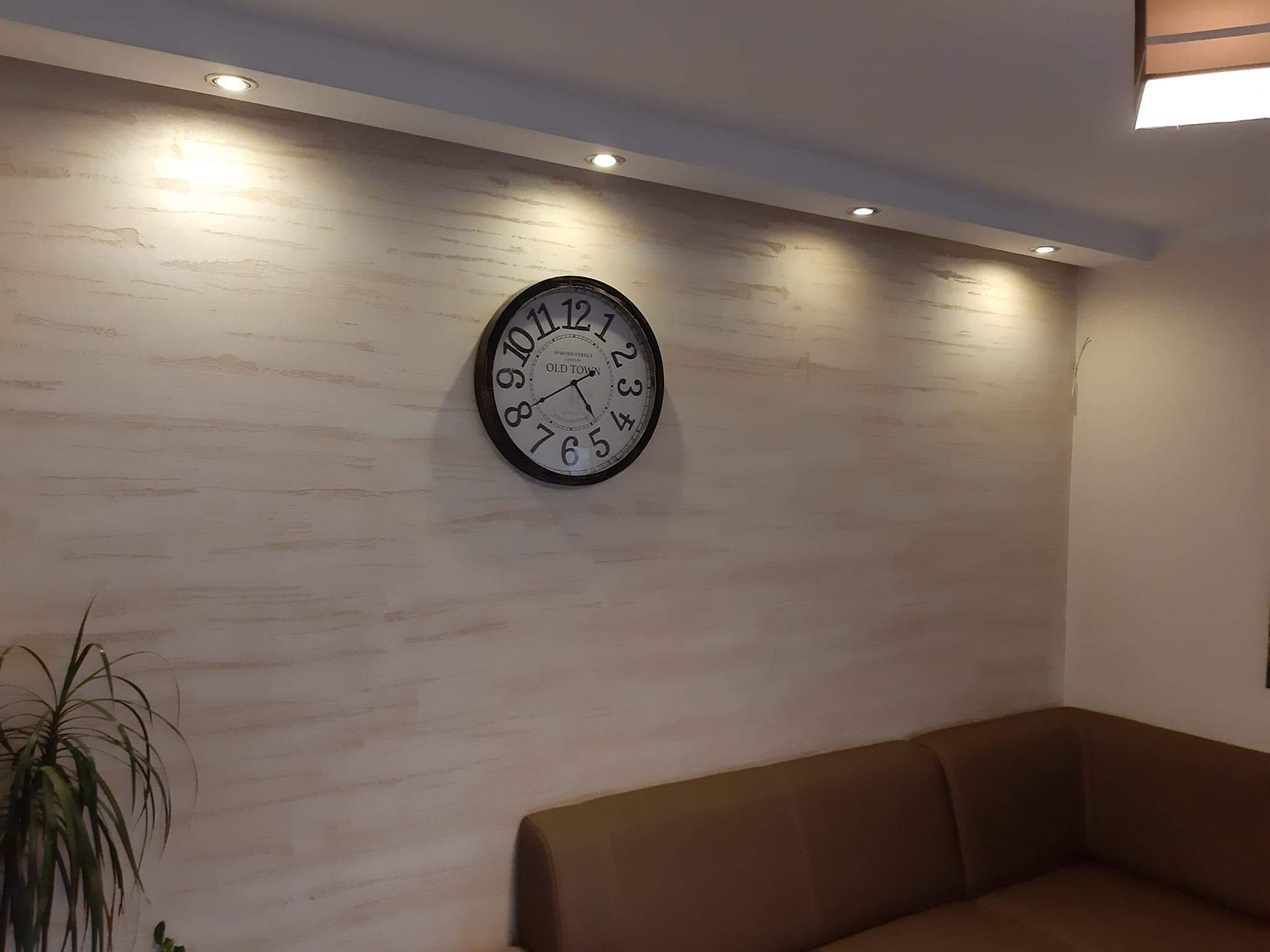 Ściana w salonie z dekoracyjną, beżowo-białą fakturą, okrągły zegar w stylu retro z napisem OLD TOWN, podwieszany sufit z oświetleniem punktowym, fragment brązowej sofy i zielona roślina doniczkowa.