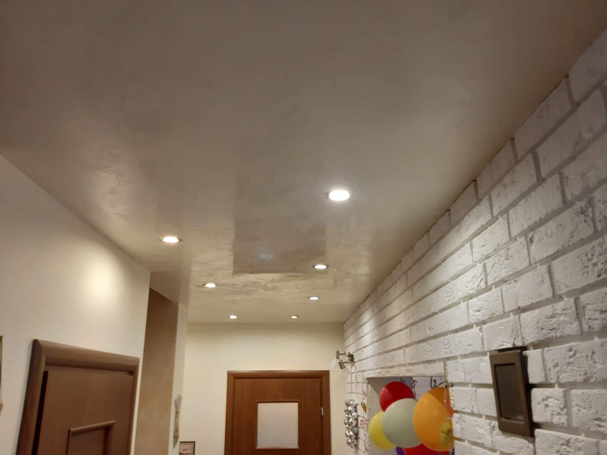 Wykończone wnętrze z dekoracyjnym tynkiem na suficie, oświetleniem punktowym i białą ścianą z cegły, widoczne drzwi i dekoracje wiszące na ścianie.