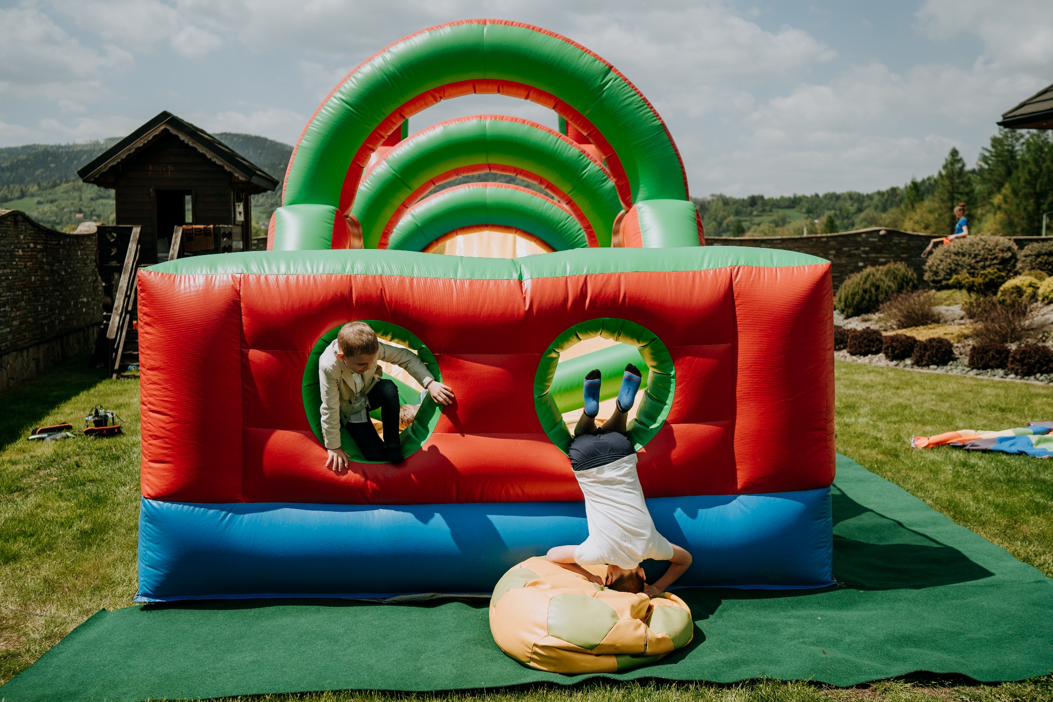 Dzieci bawią się na dmuchanym zamku z tunelem; jeden chłopiec wchodzi przez okrągły otwór, drugi stoi na głowie oparty o puf przed zamkiem.