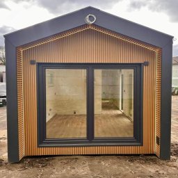 PM HOUSES Patryk Mocek - Nowoczesny, modułowy domek z drewnianą elewacją i oknami w Głogowie. Ciemna blacha na dachu, oświetlenie LED podkreśla krawędzie. Widok z zewnątrz, surowy stan.