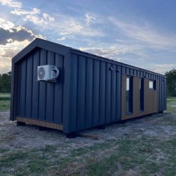 PM HOUSES Patryk Mocek - Nowoczesny, mobilny dom w ciemnej okładzinie z klimatyzacją na bocznej ścianie. Widoczny fragment samochodu i zielone otoczenie. Ujęcie z niskiej perspektywy.