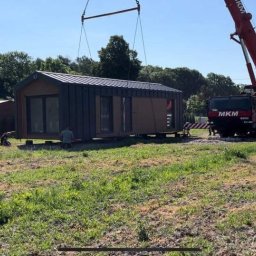 PM HOUSES Patryk Mocek - Transport modułowego domu z szarym dachem i drewnianą elewacją za pomocą czerwonego dźwigu na tle zieleni i nieba, widoczny pracownik.