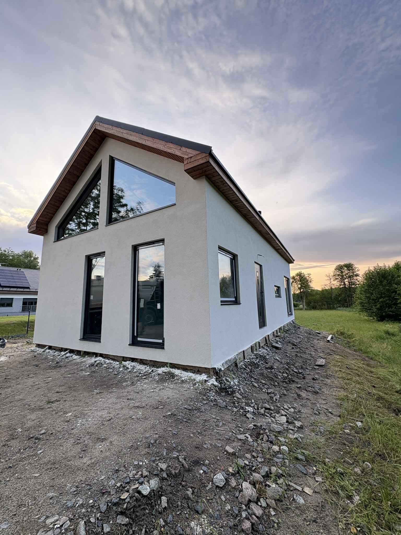 Nowoczesny dom jednorodzinny z dużymi oknami i drewnianym wykończeniem dachu, widok z zewnątrz, teren wokół domu w trakcie prac wykończeniowych.