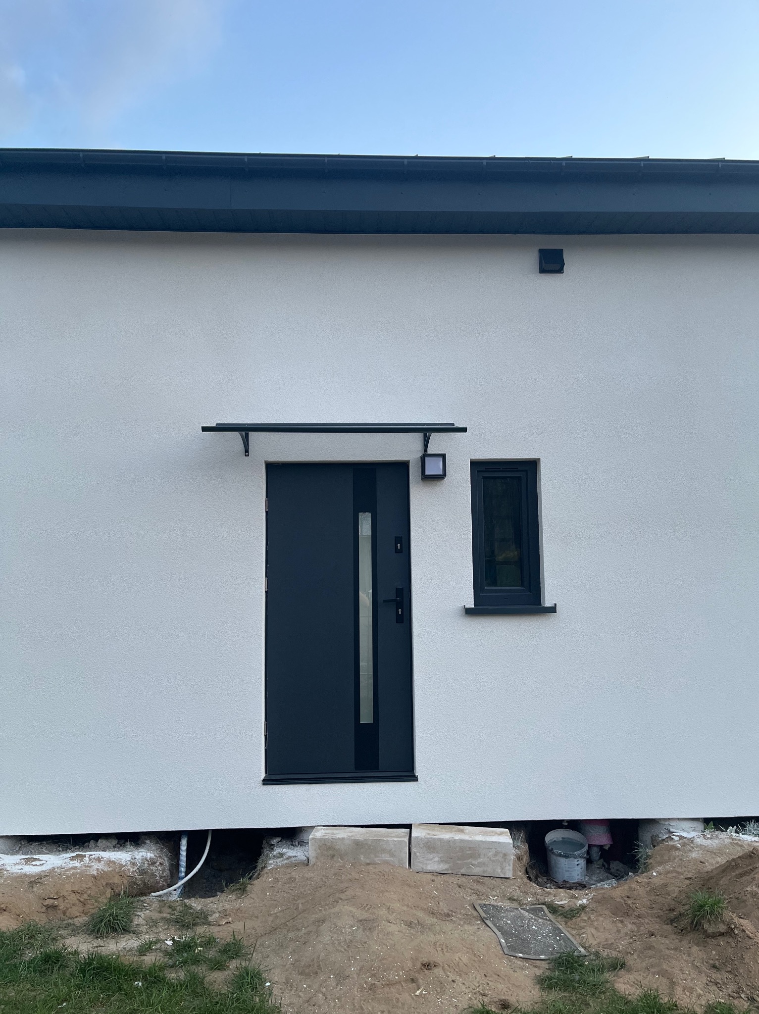 Elewacja budynku z nowoczesnymi drzwiami wejściowymi i małym oknem, widoczne wystające kable i elementy instalacji pod ścianą.