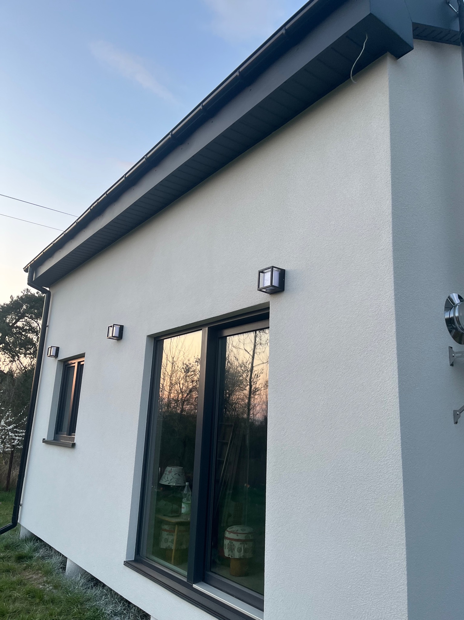 Elewacja domu jednorodzinnego z oknami, oświetleniem zewnętrznym i fragmentem rury wentylacyjnej. Ściana w jasnym odcieniu, okna w ciemnych ramach. Ujęcie z dołu, widoczne niebo.