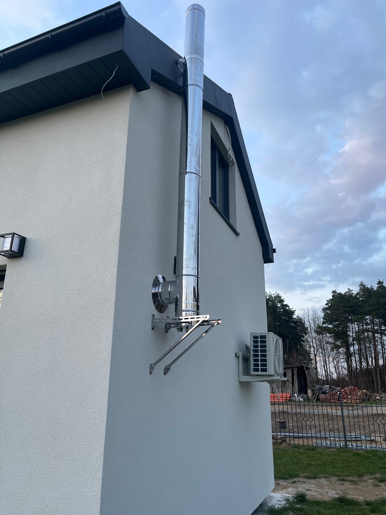 Zewnętrzny, stalowy komin zamontowany na ścianie domu z klimatyzatorem. Widoczne elementy montażowe i fragment instalacji. Tło stanowią drzewa i błękitne niebo.